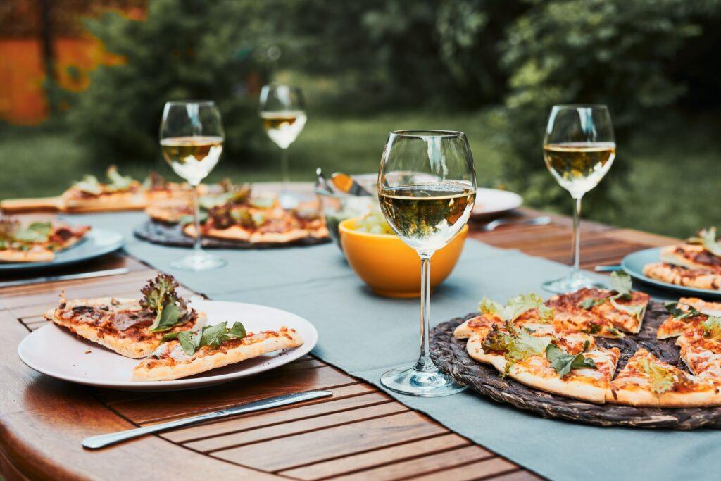 Pizzastücke und Weingläser auf einem gedeckten Gartentisch. Die Pizza wurde frisch im Pizzaofen zubereitet und ist bereit, bei einem gemütlichen Gartendinner genossen zu werden.

