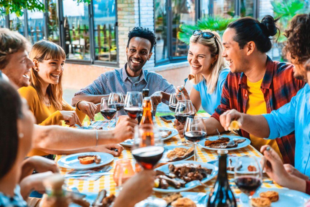 Freunde genießen ein gemeinsames Gartendinner bei sonnigem Wetter. Lachen und gute Gespräche begleiten das Essen, das frisch im Pizzaofen zubereitet wurde.