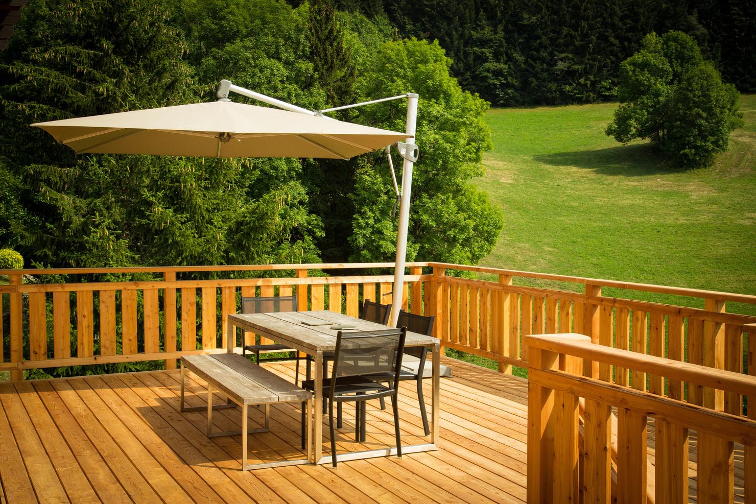 Eine Holzterrasse mit einem rustikalen Esstisch und zwei Bänken sowie zwei Stühlen. Über dem Tisch hängt ein großer, cremefarbener Sonnenschirm, der Schatten spendet. Die Terrasse bietet eine malerische Aussicht auf grüne Wiesen und dichte Wälder im Hintergrund. Die warmen Holztöne der Terrasse harmonieren mit der natürlichen Umgebung, und die Szene strahlt Ruhe und Entspannung in einer ländlichen Umgebung aus.