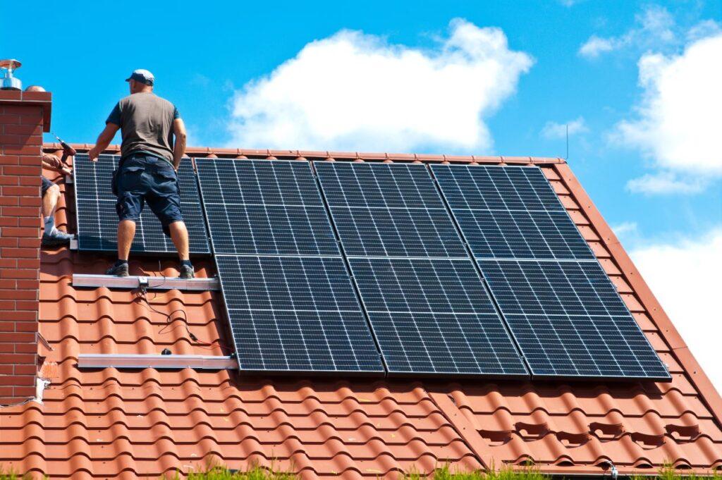 Handwerker installieren Solarmodule und einen Photovoltaik-Wechselrichter auf einem roten Ziegeldach unter blauem Himmel