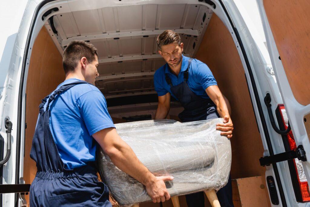 Zwei Männer laden ein mit Stretchfolie umwickeltes Möbelstück in einen Lieferwagen für einen sicheren Transport