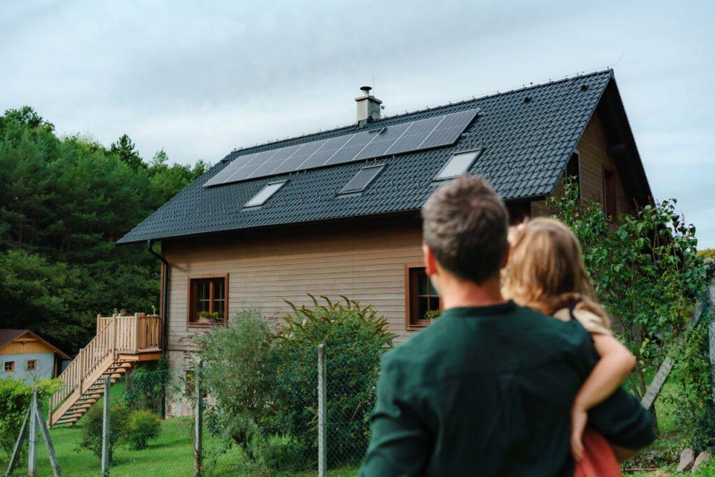 Ein Mann mit Kind betrachtet ein modernes Holzhaus mit schwarzem Dach und installierter Photovoltaikanlage, umgeben von Natur – nachhaltige Energieversorgung in Gera.